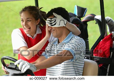 Stock Photo - Female golfer covering the eyes of her partner as he drives a golf cart. Fotosearch