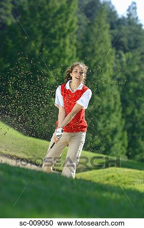 Stock Image - Sand explosion as a female golfer plays a bunker shot. Fotosearch