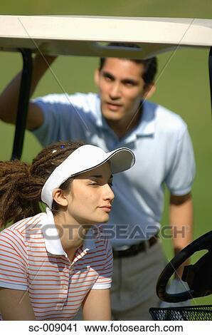 Stock Image - Side view of a woman sitting in a golf cart while a man is seen at the back. Fotosearch