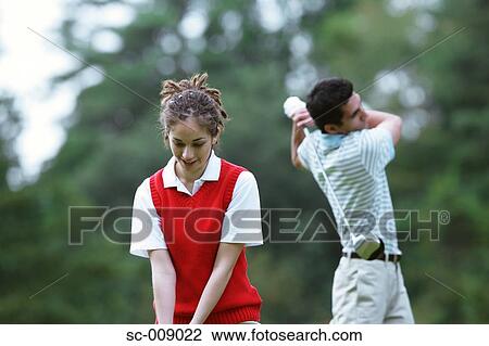 Stock Image - Woman smiling as a man plays a golf stroke in the background. Fotosearch