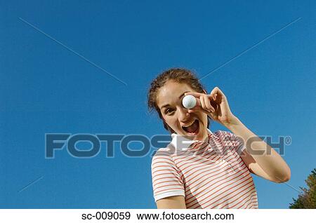 Stock Photo - Woman smiling at the camera as she holds a golf ball in front of her left eye. Fotosearch