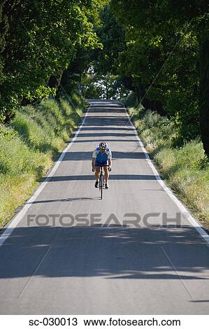 Stock Image - Cyclist riding bicycle on deserted road, front view. Fotosearch