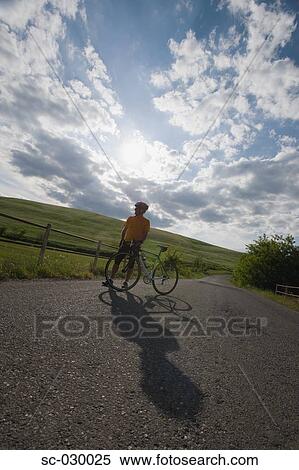 Stock Photography - Cyclist standing with bike on road beneath cloudy sky. Fotosearch