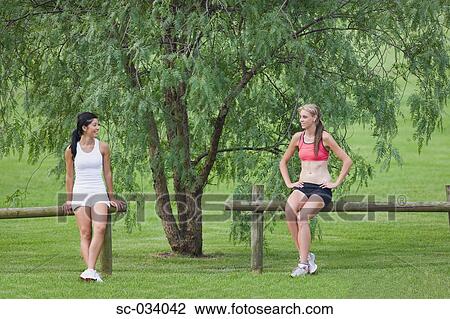Stock Image - Two young women sitting on log near tree. Fotosearch