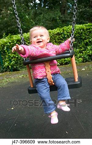 Stock Photo - 1 year old girl sitting in a swing, smiling and pointing off camera. Fotosearch