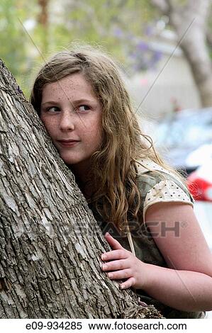 Stock Photography - 13-year-old girl with blonde-brown long hair and freckles, half leaning against, half hiding behind a tree, up-to-something expression. Fotosearch