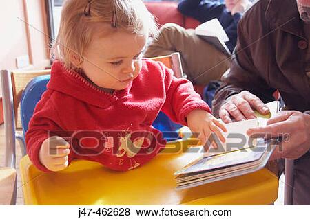 Stock Photo - 18 month old girl in a cafe looking at a book with her grandad. Fotosearch