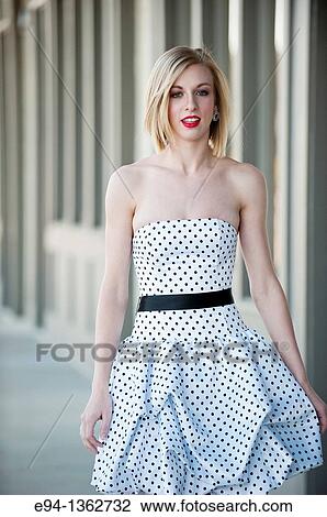 Stock Image - 19 year old blond woman walking on a sidewalk in a small town wearing dress. Fotosearch