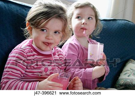 Stock Image - 2, 3 year old girls drinking juice through a straw, looking off camera, smiling. Fotosearch