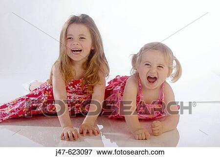 Stock Photo - 2 little girl, aged 2 and 4, lying on their tummies in the studio, laughing. Fotosearch