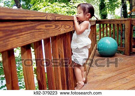 Stock Image - 2-year-old Vietnamese girl looking over the railing of a deck. Fotosearch