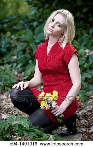 Stock Photograph - 21 year old blond woman wearing a red sweater and black boots and stockings holding a bouquet of yellow flowers on a garden path looking at camera.. Fotosearch