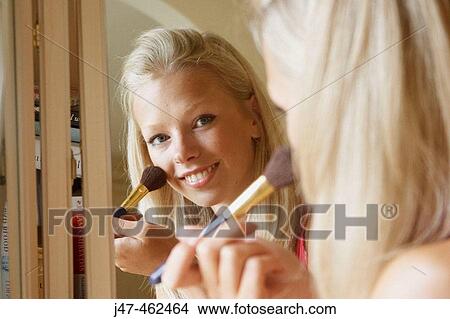 Picture - 21 year old girl looking at the camera through a mirror putting on her make up.. Fotosearch