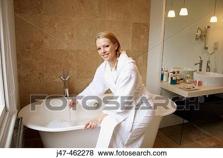 Stock Photo - 21 year old girl sitting on the edge of the bath, smiling, holding her hand under the water.. Fotosearch