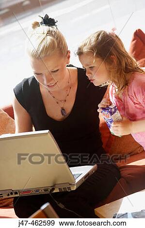 Stock Photo - 21 year old girl working on a laptop on her knee, with a 3 year old girl watching. Fotosearch