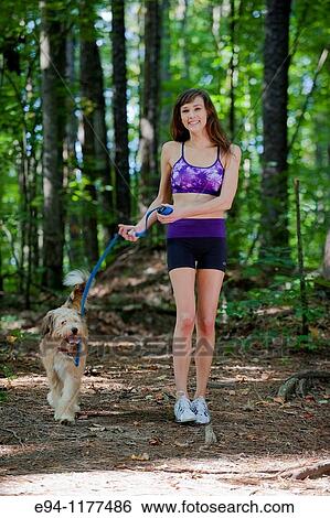 Stock Photograph - 23 year old brunette woman dressed in exercise clothing walking her dog through a forest. Fotosearch