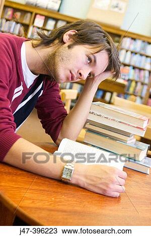 Stock Image - 24 year old man leaning over a book, head in hand, in the library. Fotosearch