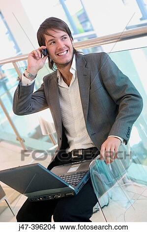 24 year old man sitting in chair talking on mobile phone, smiling, working on laptop View Large Photo Image Picture - 24 year old man sitting in chair talking on mobile phone, smiling, working on laptop. Fotosearch