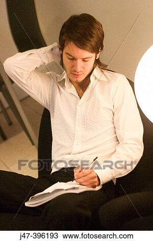 Stock Image - 24 year old man sitting in chair writing, one arm behind head. Fotosearch