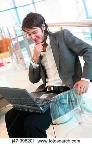Stock Image - 24 year old man sitting in chair, working on laptop, smiling, hand to chin. Fotosearch