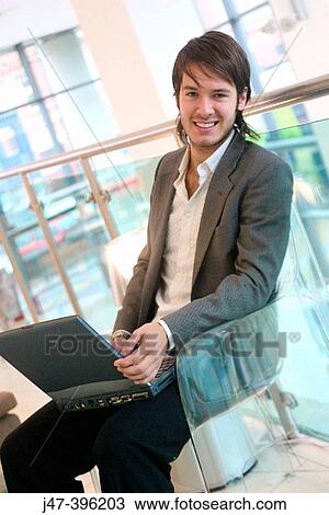 Stock Image - 24 year old man sitting in chair, smiling into camera, working on laptop. Fotosearch