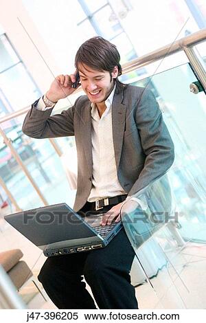 Stock Photography - 24 year old man sitting in chair talking on mobile phone, working on laptop. Fotosearch
