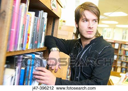 Stock Image - 24 year old man standing, in the library, pulling a book off the shelf. Fotosearch