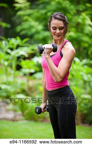 Stock Image - 25 year old brunette woman in a garden settng wearing work out clothing with dumbbells. Fotosearch