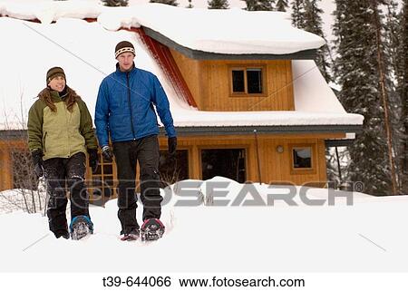Stock Photograph - 25_35 year old couple walking on snowshoes with a winter lodge in the background.. Fotosearch