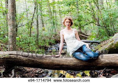 Picture - 29 year old blond woman in a forest sitting a on log wearing blue jeans and a white top. Fotosearch