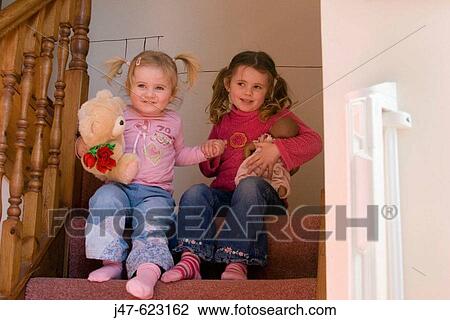 Stock Image - 3 and 2 year old sisters sitting on the stairs holding hands, smiling.. Fotosearch