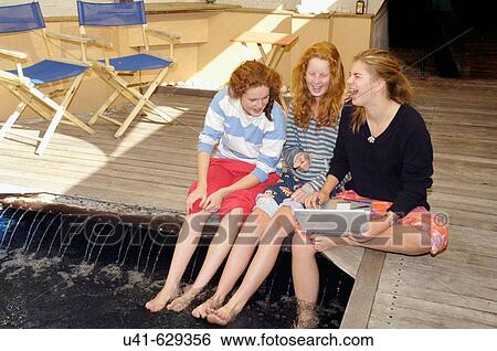 Stock Photograph - 3 friends sitting by swimming pool while checking internet on laptop. Fotosearch