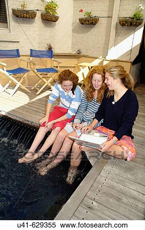 Stock Photography - 3 friends sitting by swimming pool while checking internet on laptop. Fotosearch