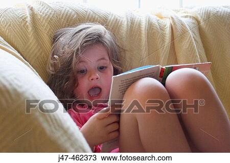 Stock Image - 3 year old girl on the settee, with her knees up reading a bedtime story mouth wide open with surprise. Fotosearch