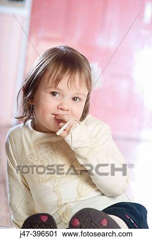 Stock Image - 3 year old girl sitting on the floor, smiling into camera, hand to mouth. Fotosearch