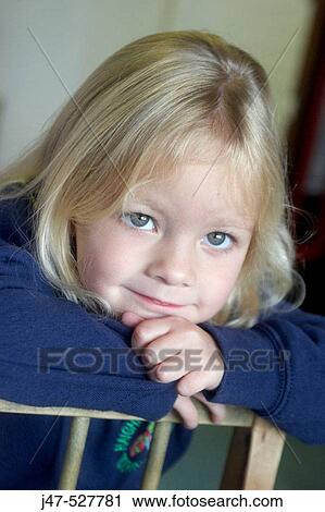 Stock Image - 3 year old girl smiling into camera at nursery, leaning onto her hands over the back of a chair. Fotosearch