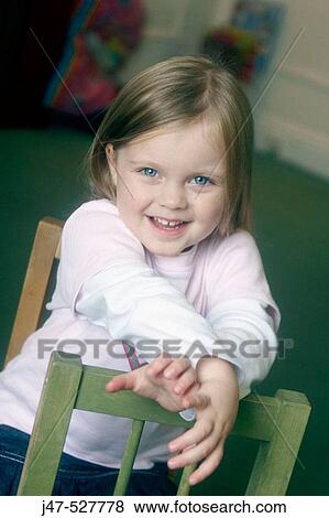 Stock Photo - 3 year old girl smiling into camera at nursery, leaning the back of a chair. Fotosearch