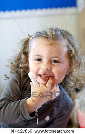 Stock Photograph - 3 year old girl smiling off camera at nursery, hand up to her mouth. Fotosearch