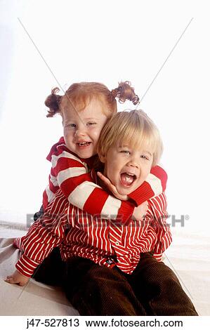 Stock Image - 3 year old girl with red hair cuddling her brother, laughing and smiling into camera, in the studio. Fotosearch