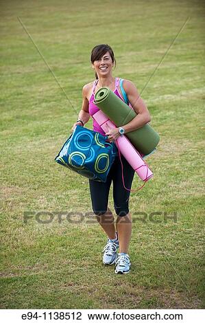 Stock Image - 30 year old woman in work out clothing walking on grass carrying yoga matts and work out equipment. Fotosearch