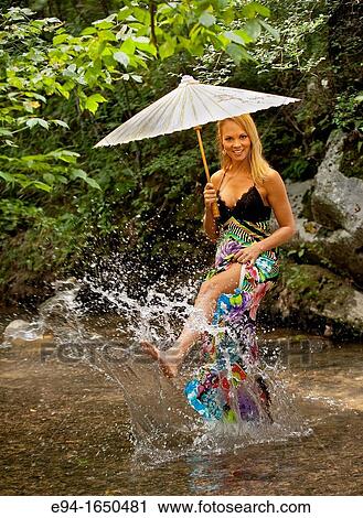 Stock Image - 34 year old blond woman in a long flower print dress standing in small creek looking over her shoulder at the camera. Fotosearch