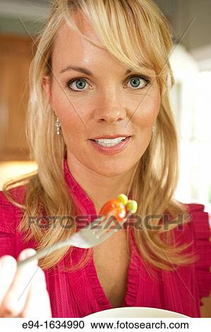 Stock Image - 35 year old blond woman standing in a kitchen with a salad holding a fork, eyes to camera. Fotosearch