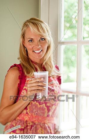 Stock Photo - 35 year old blond woman standing in a kitchen holding cold fruit drink. Fotosearch