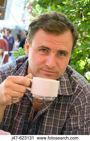 Stock Image - 37 year old man sitting drinking coffee, outside, smiling just off camera. Fotosearch