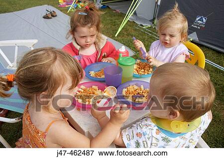 Stock Photo - 4 children camping, sitting round a table eating dinner, one is feeding the little baby.. Fotosearch