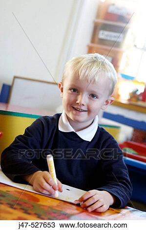 Stock Image - 4 year old boy smiling into camera at nursery, drawing. Fotosearch