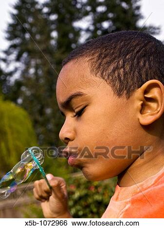 Stock Photograph - 5 yr old multiracial boy blowing soap bubbles through soap wand side view, Vertical. Fotosearch