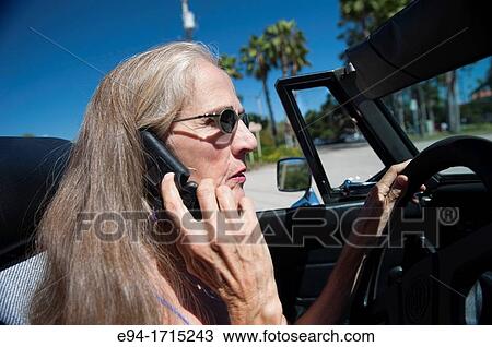Stock Image - 57 year old woman with long hair driving her MG convertible car and talking on a cell phone. Fotosearch