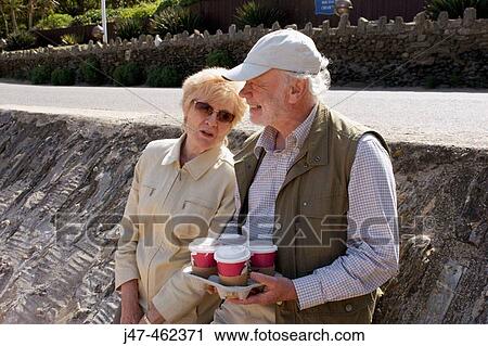 Stock Image - 70 year old man and wife leaning on a wall at the seaside with takeaway coffee, laughing. Fotosearch