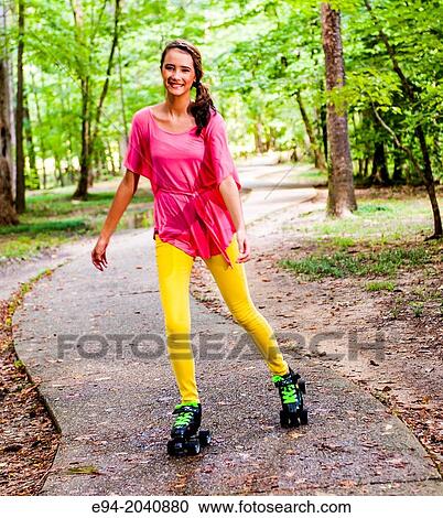 Stock Image - A 14 year old brunette girl on roller skates weaing yellow jeans and a pink top in a park setting.. Fotosearch
